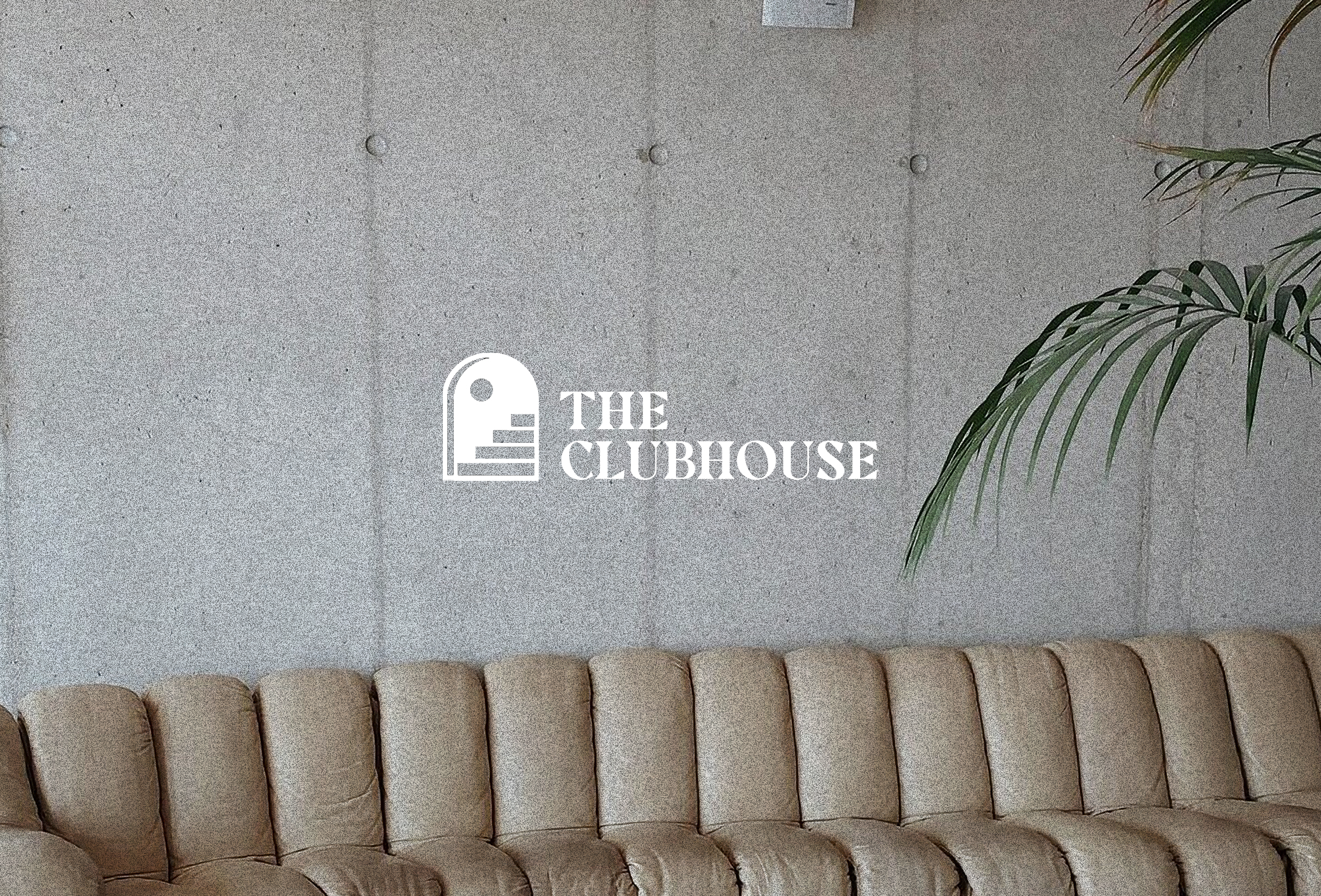 An image of The Clubhouse lounge area including a long beige leather couch leaning against a grey concrete wall and a palm tree off to the right side. A white Clubhouse logo is centered on top of the image