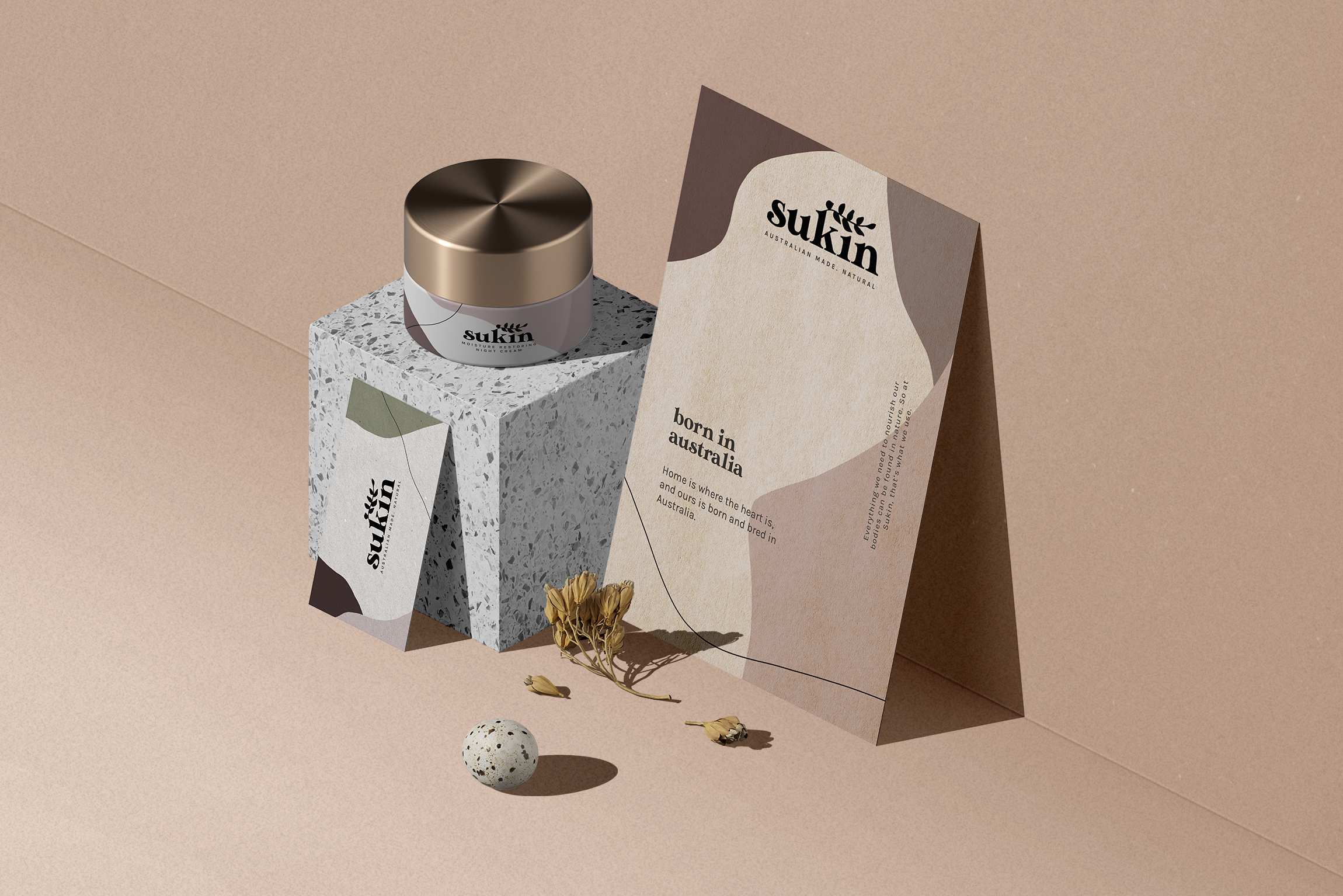 A display that is showcasing a Sukin business card leaning on a terrazzo patterned cube and a Sukin postcard slanted on a wall beside it. On top of the cube is a tubbed moisturizer with a gold lid.